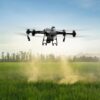An industrial Agriculture Spraying Drone flying over a vibrant green crop field delivering precision liquid pesticide treatment via automated downward spray nozzles.