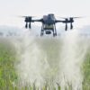 The commercial Dji Agras t25 multirotor spray drone hovering steadily above a green crop field while releasing a dense liquid mist from its dual atomizing sprinklers.